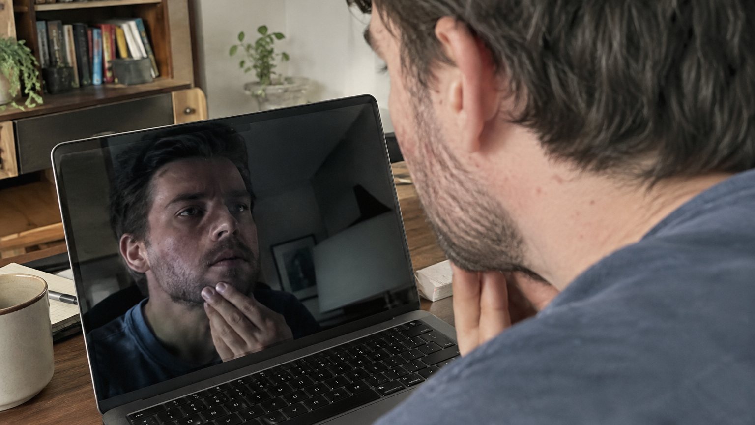 An adult man with jawline breakouts checks his laptop reflection before a video meeting.