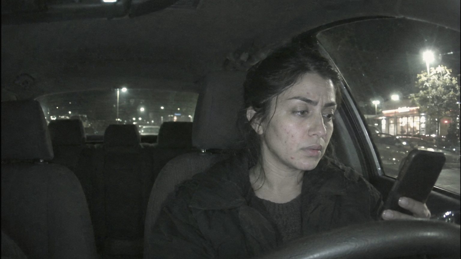 A worried adult woman checks visible jawline acne in a dark phone screen inside a parked car before a date
