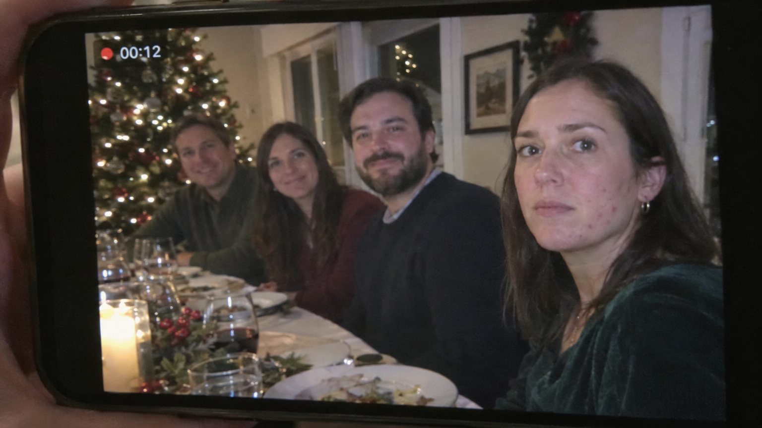 An adult woman with ordinary visible acne stays calm while someone records a holiday group photo on a phone.