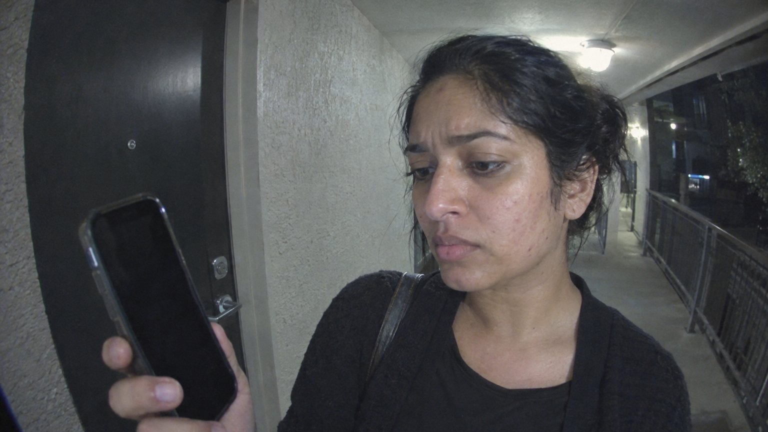 A South Asian adult woman checks visible acne in a dark phone screen at an apartment door before going out.